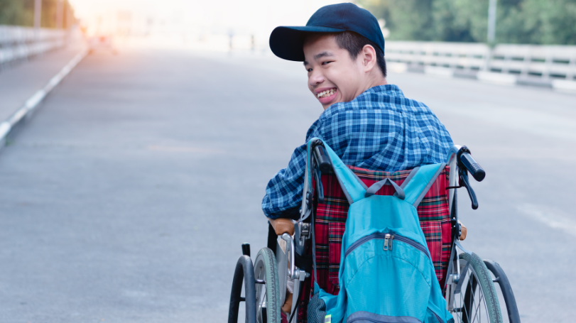 A young person using a wheelchair with a backpack mounted on the back of the chair while outdoors.