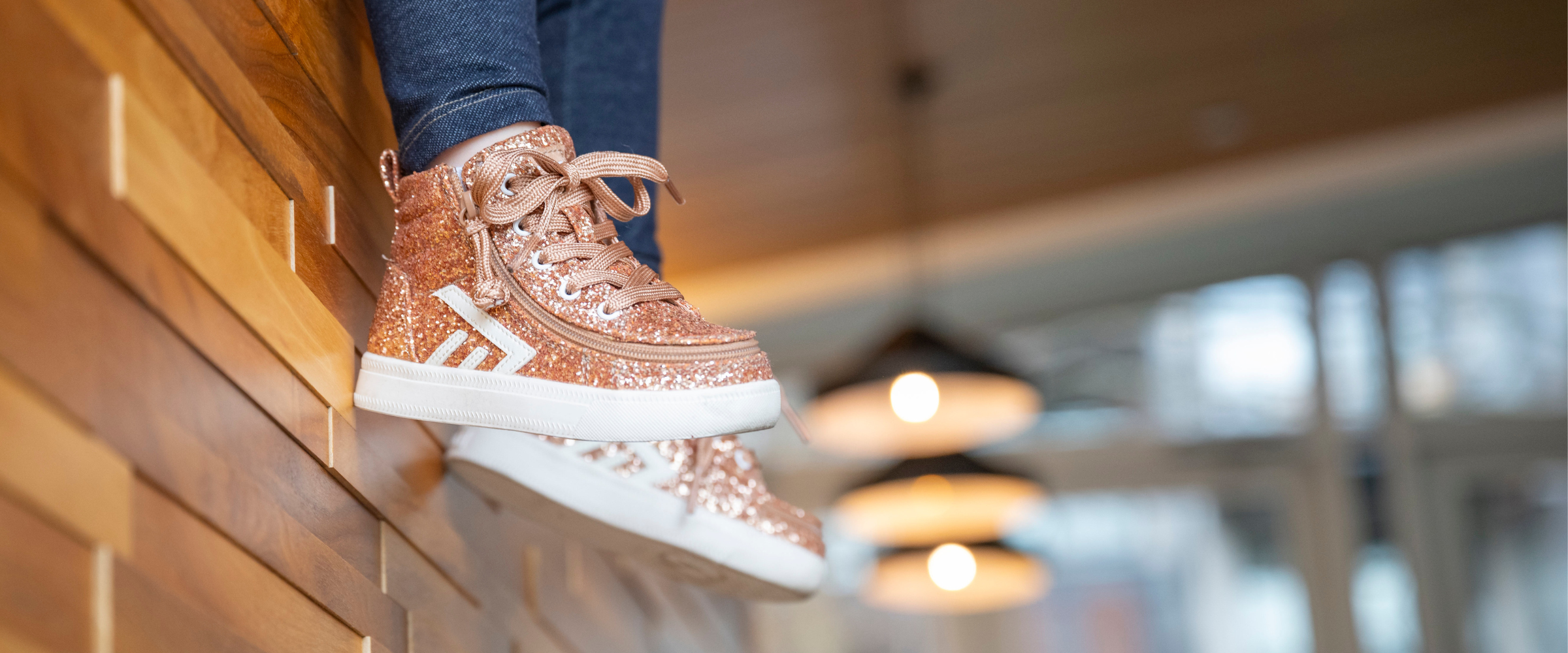 Rose gold glitter Billy Footwear adaptive high-top sneakers with side zip closure, shown dangling over a wooden ledge in a warm interior setting