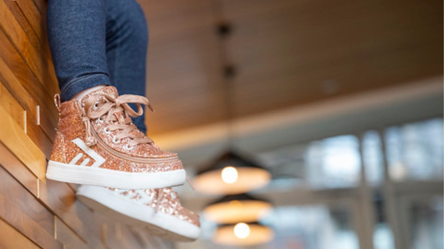 Kid wearing rose gold sparkly BILLY classic high top adaptive sneakers, sitting on a counter in a restaurant. 