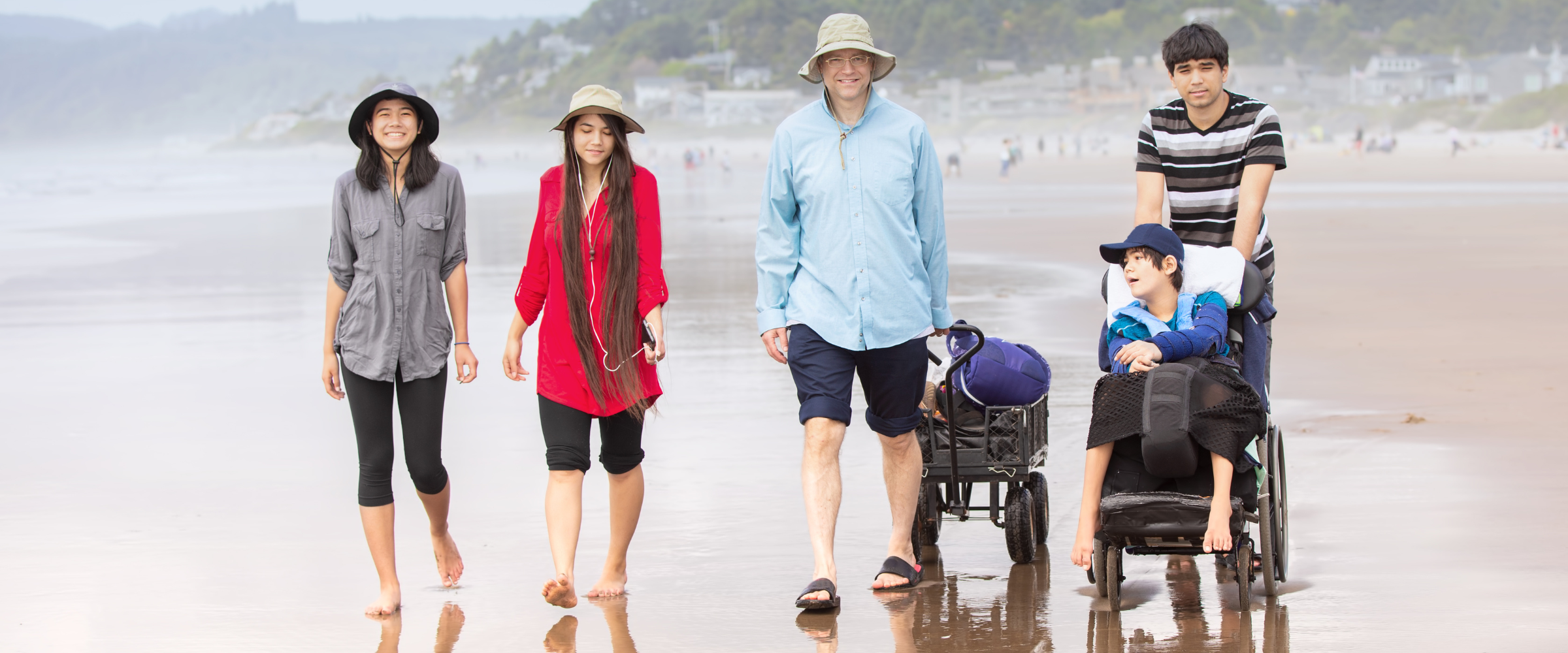 A family walking along a beach shoreline. Two women, a man pulling a wagon, and a child pushing a younger child in a wheelchair.