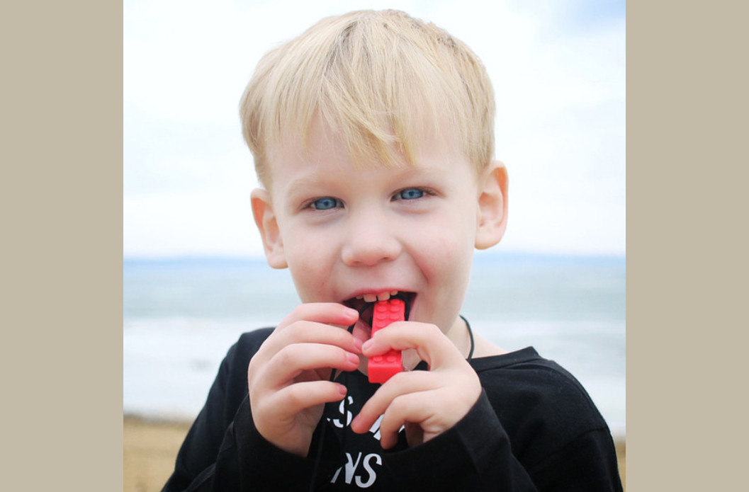 Young boy chewing on a red brick-textured sensory chew necklace pendant at the beach