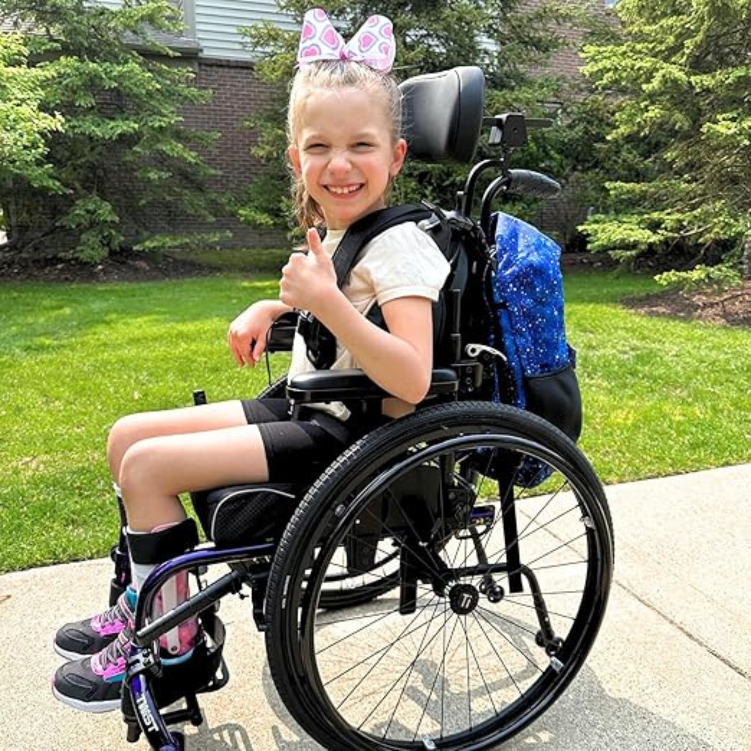 A child using a wheelchair with a backpack mounted on the back of the chair outdoors.