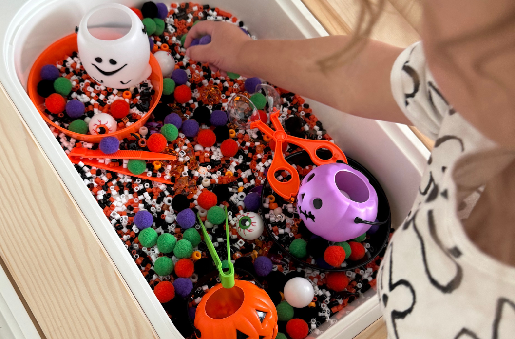 Child reaching into a Halloween-themed sensory bin filled with beads, pom poms, scooping tools, and festive containers.