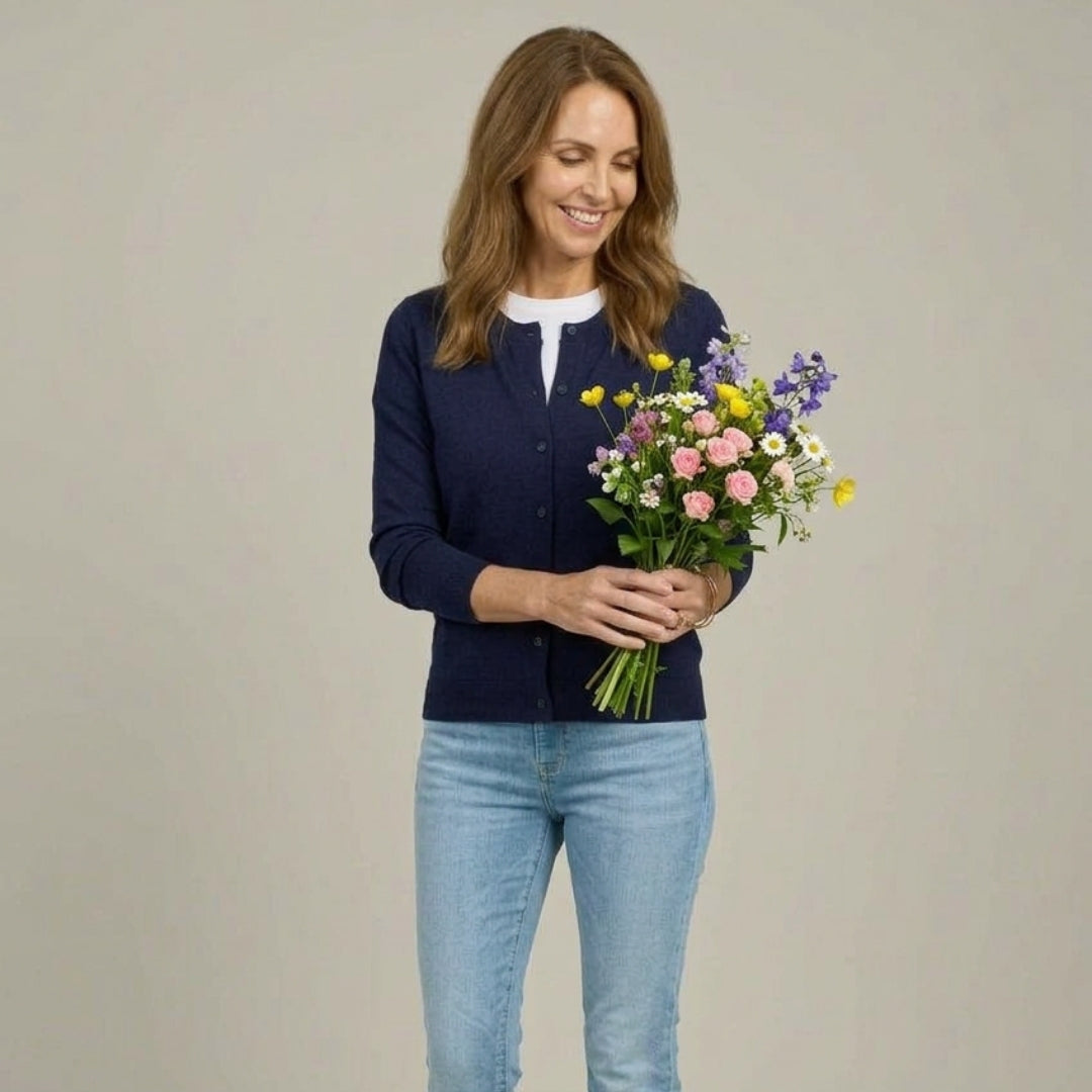 A woman wearing MagnaReady navy magnetic knit cardigan over a white top and light blue jeans, holding a colorful bouquet of wildflowers against a plain beige studio background.
