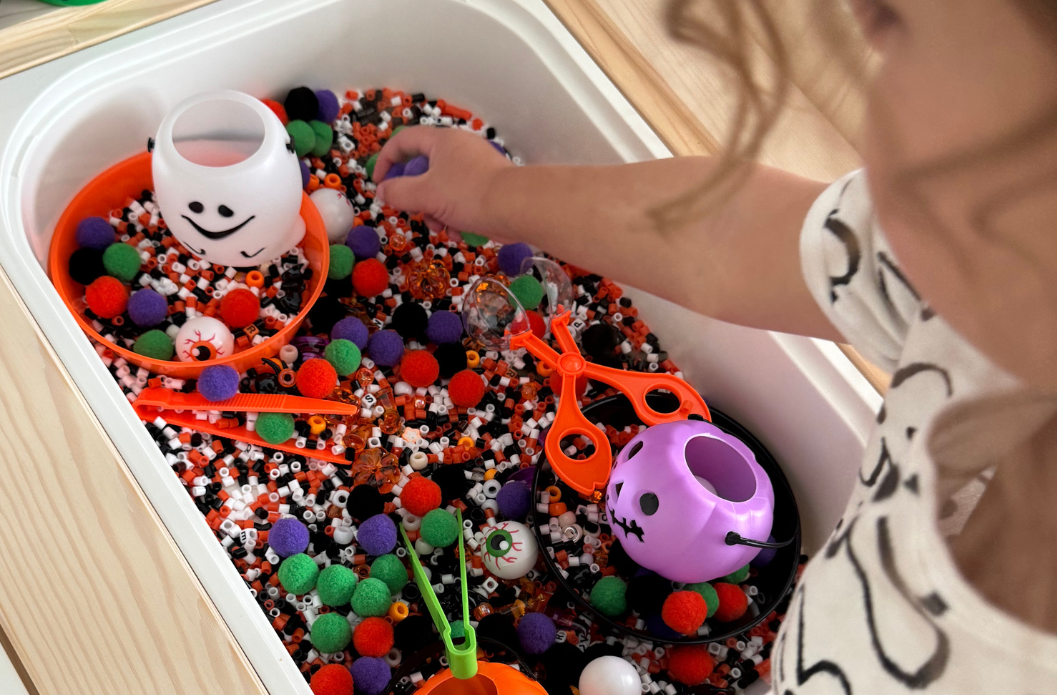 Child playing with a DIY Halloween sensory bin filled with pom poms, scoops, and sensory materials for hands-on Halloween play