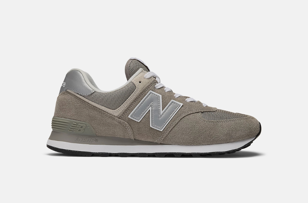 A single New Balance 574 sneaker in a taupe-gray color, a supportive athletic shoe commonly recommended for orthotics and SMOs, featuring a suede and mesh upper, a large 'N' logo on the side, and a white midsole. 