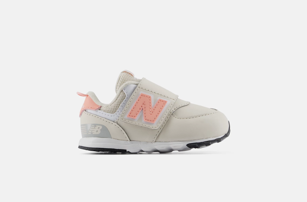 A New Balance 574 sneaker for children, featuring a light beige upper with a coral pink 'N' logo, a hook-and-loop strap closure for easier on and off, and a white and black sole, shown against a light gray background.