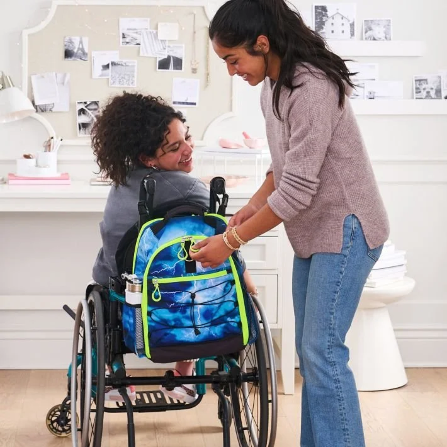 Teen girl in a wheelchair smiling as a friend zips up her blue lightning-print adaptive backpack, mounted to the back of her wheelchair
