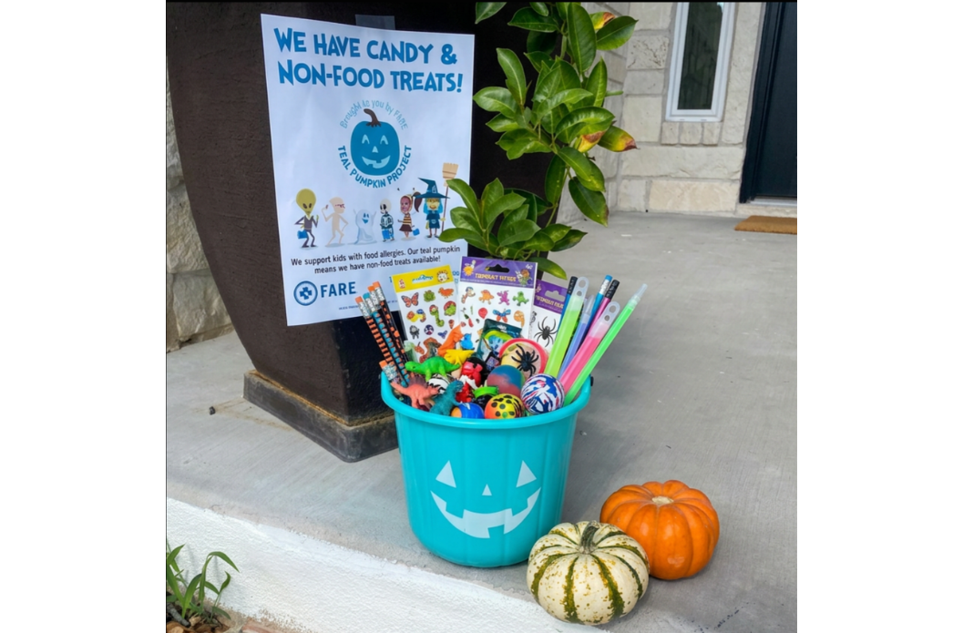 A teal bucket filled with Halloween-themed non-candy treats like stickers, glow sticks, and pencils, sitting on a concrete porch next to a small orange pumpkin and a striped gourd and a sign that says, "We have candy & non-food treats!"