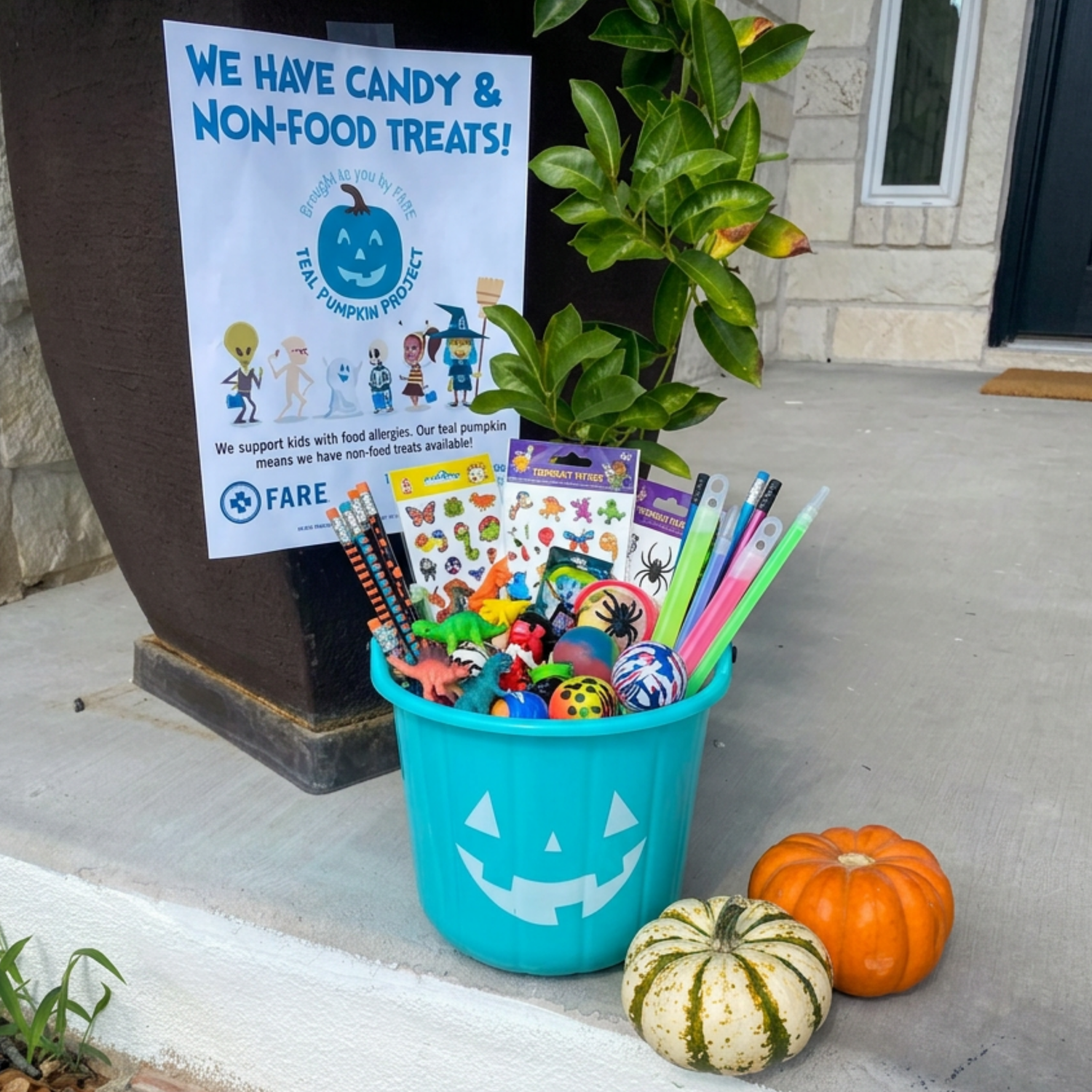 A Teal Pumpkin Halloween treat basket filled with non-candy treats like stickers, pencils, glow sticks, and toys, placed on a front porch next to a sign reading 'We Have Candy & Non-Food Treats!' and a small jack-o'-lantern and a striped gourd.