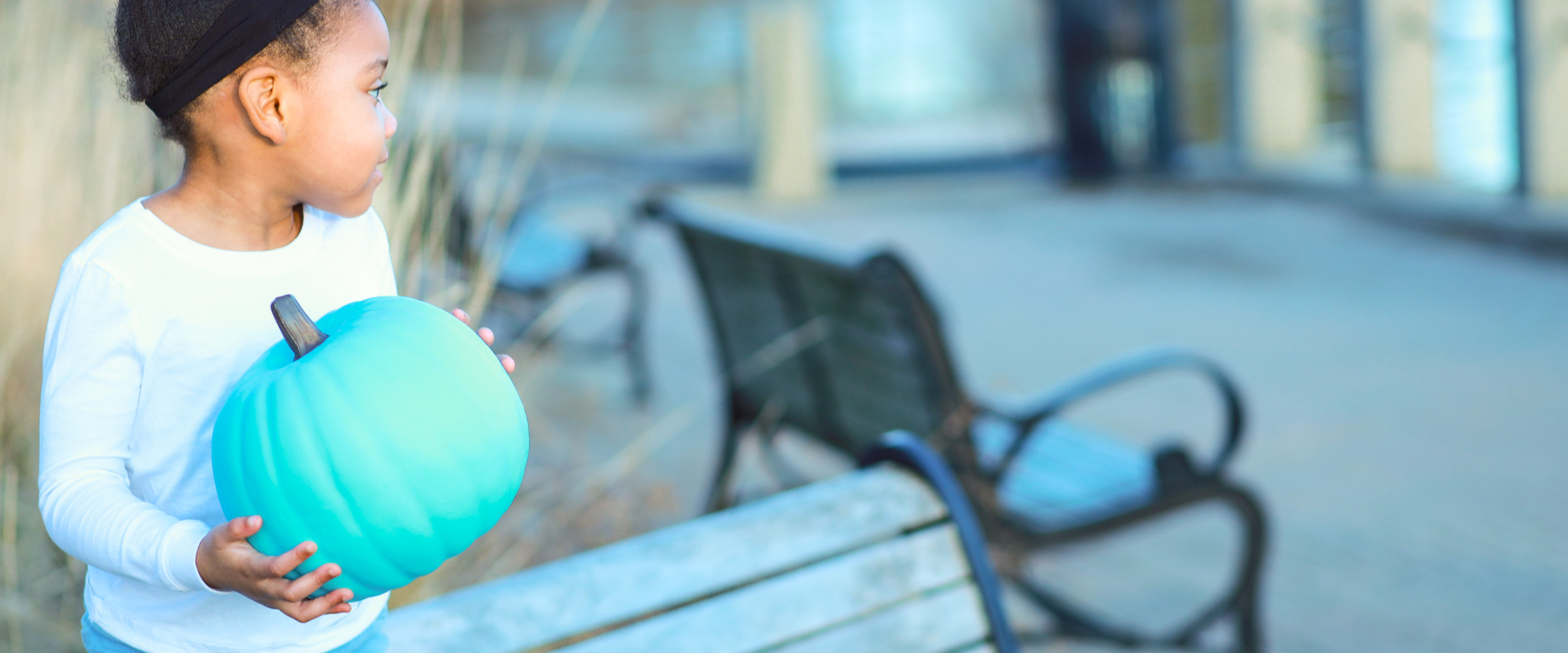A young child wearing a white long-sleeve shirt holds a bright teal decorative pumpkin outdoors next to a wooden bench.