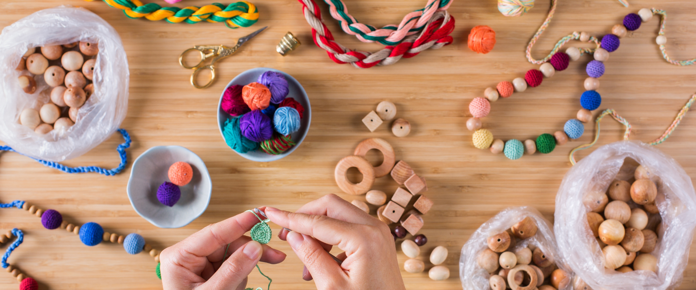 Hands crocheting a small bead surrounded by wooden beads, colorful yarn balls, braided rope sensory necklaces, and chewelry-making supplies spread across a wooden table, overhead view
