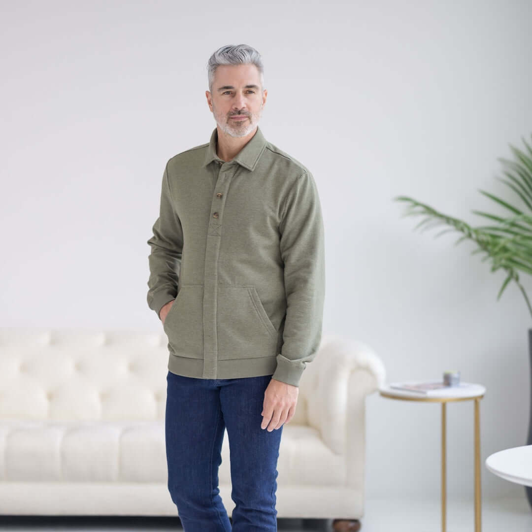 Man wearing MagnaReady heathered olive magnetic front sweatshirt, full front view, standing in a living room with a white couch and plant.