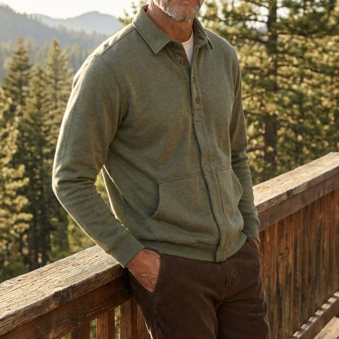 Man wearing a heathered olive magnetic front sweatshirt and brown pants standing on a wooden deck with trees in the background.