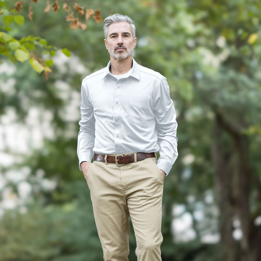 A man wearing a MagnaReady white long-sleeve, magnetic button down cooling shirt with khaki pants, and a brown belt, standing outdoors in a park with blurred green trees in the background.