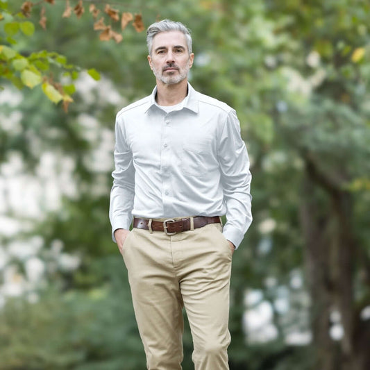 A man wearing a MagnaReady white long-sleeve, magnetic button down cooling shirt with khaki pants, and a brown belt, standing outdoors in a park with blurred green trees in the background.