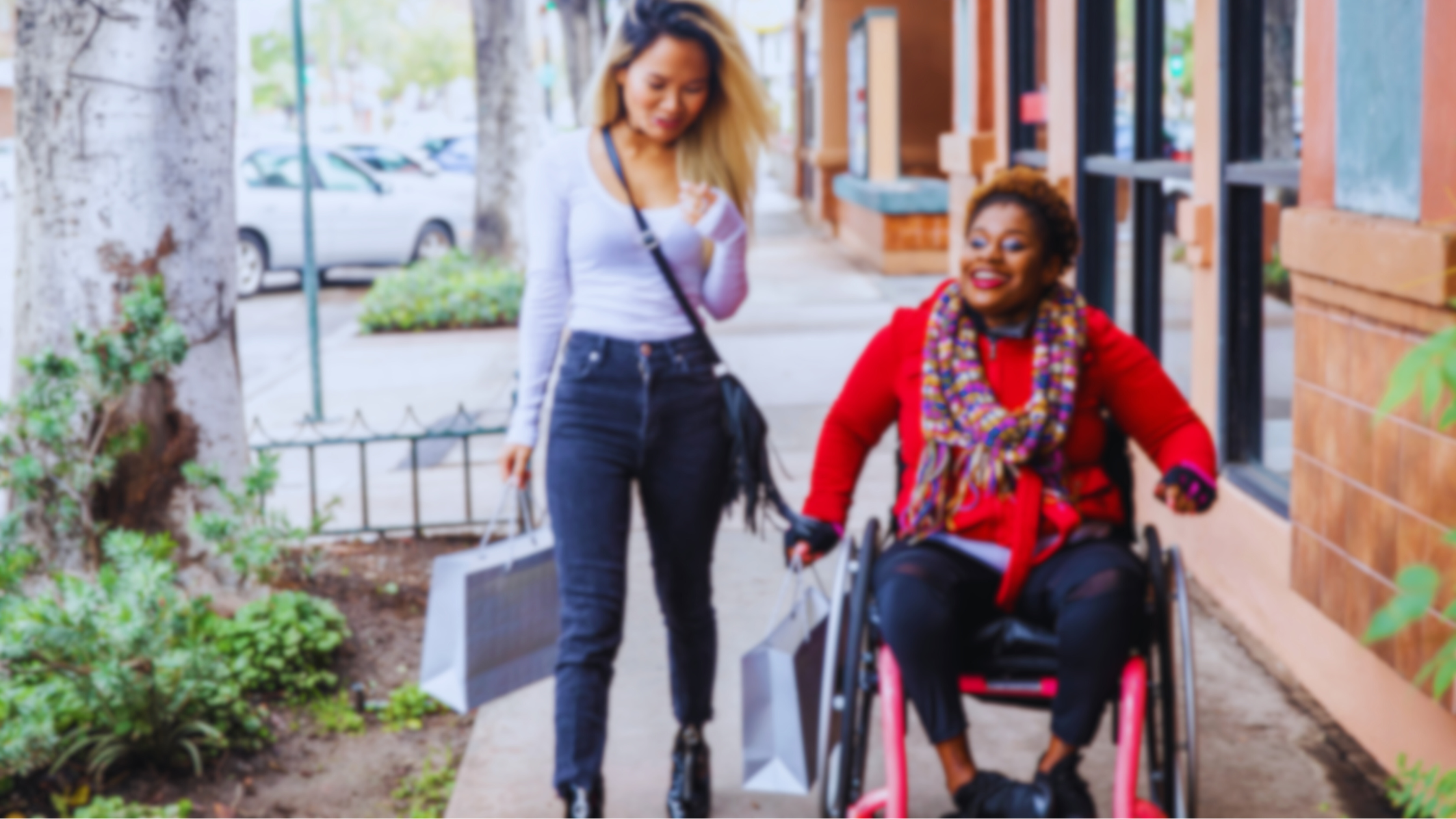 Two women holding shopping bags walking down a city sidewalk, one using a wheelchair.
