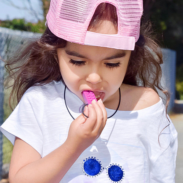 Young girl chewing on a pink brick-textured sensory chew necklace pendant outdoors