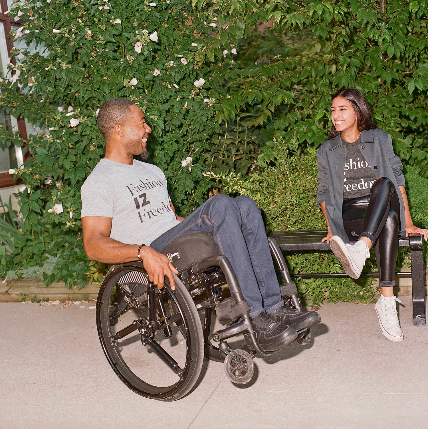 Two people wearing IZ Adaptive "Fashion iz Freedom" graphic tees laughing together outdoors — one seated in a manual wheelchair, one sitting on a bench