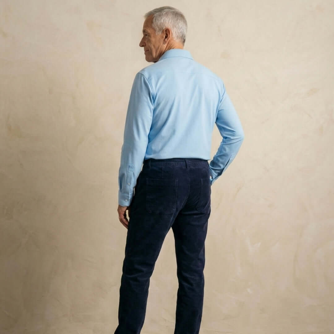 Back view of a man wearing men's navy magnetic corduroy pants and a light blue shirt against a beige wall.