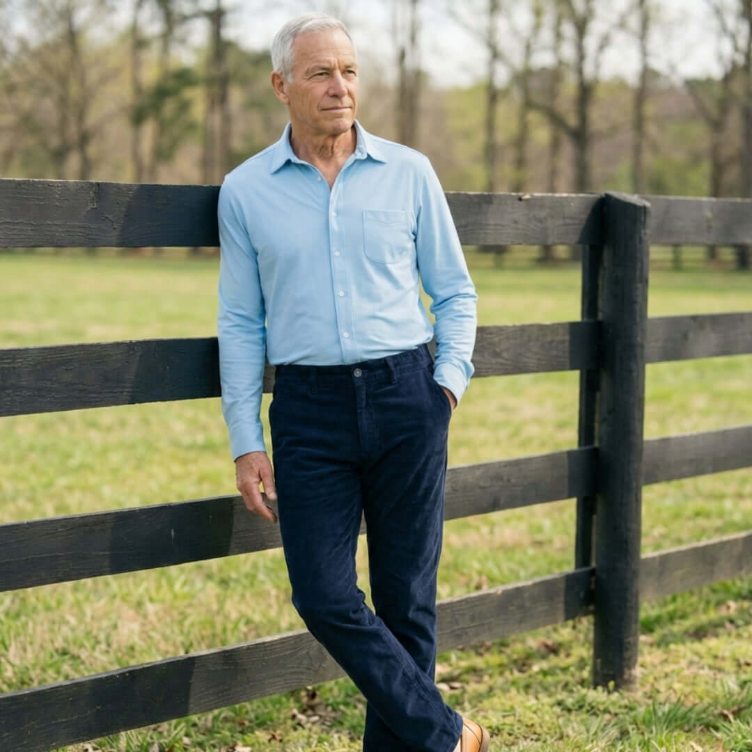 Man wearing navy magnetic corduroy pants and a light blue shirt standing by a wooden fence in a grassy field.