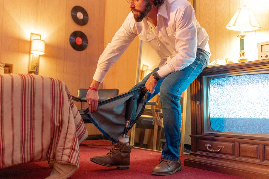 Man pulling on No Limbits adaptive jeans over a prosthetic leg in a warmly lit retro motel room