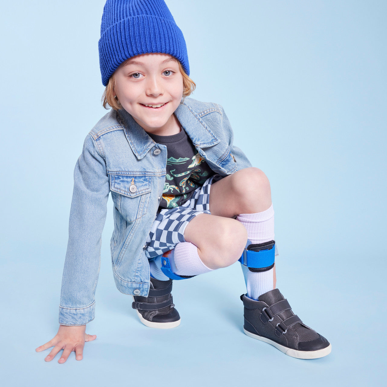 Child wearing See Kai Run adaptive high-top sneakers with AFO braces and tall white socks. He has on shorts, a black graphic tee, jean jacket, and blue beanie.