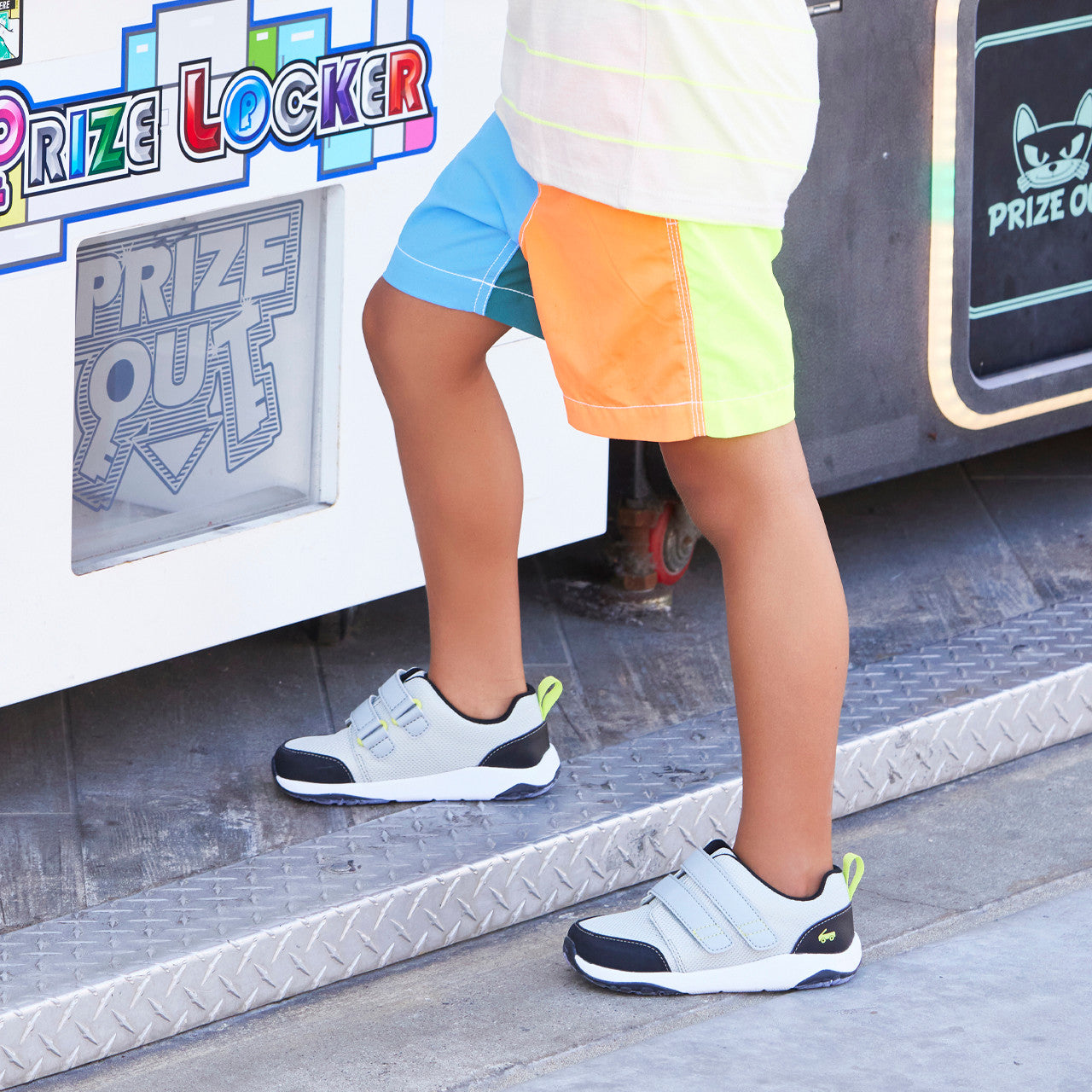 Child wearing See Kai Run Ryder Adapt adaptive sneakers in light grey. He is also wearing neon shorts standing next to a Prize Locker outside. 