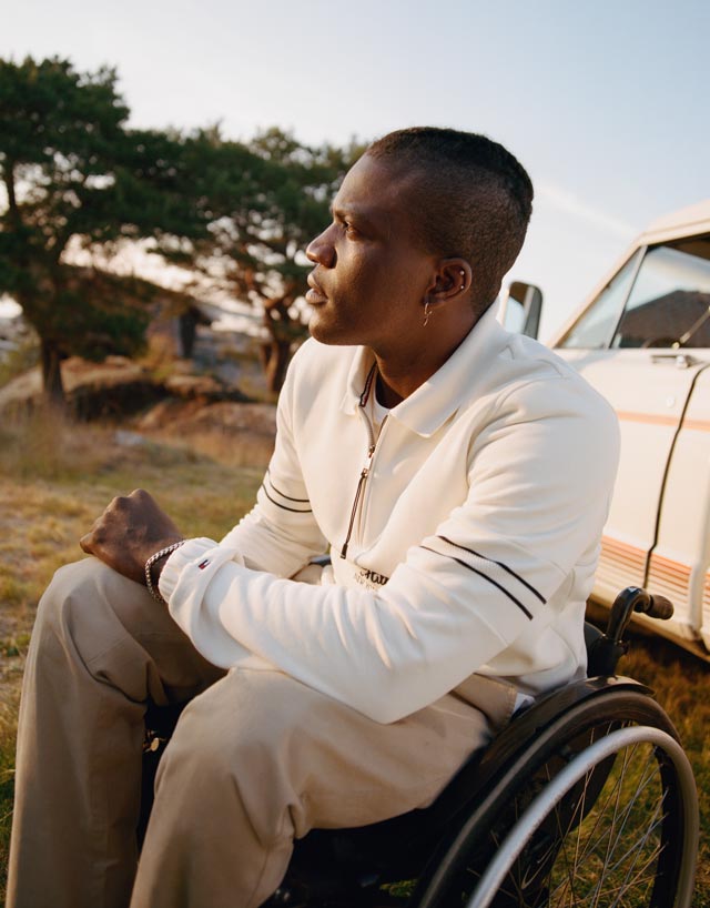 Man using a wheelchair wearing Tommy Hilfiger adaptive clothing designed for seated comfort, outside at dusk in front of a white car. 