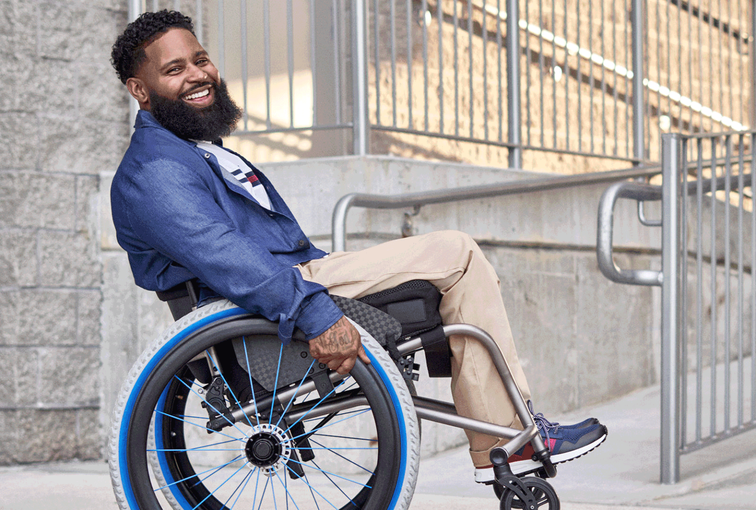 Man using a wheelchair smiling outdoors near a building entrance, wearing Tommy Hilfiger Adaptive clothing designed for seated comfort and easier dressing.