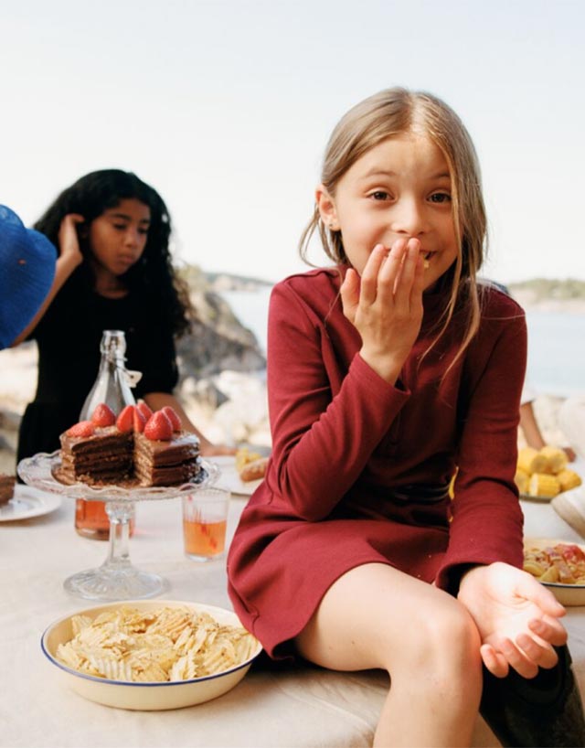 Young girl in a sensory-friendly adaptive red dress with soft fabrics and flat seams sitting at a table with food, smiling and holding her hand to her mouth.